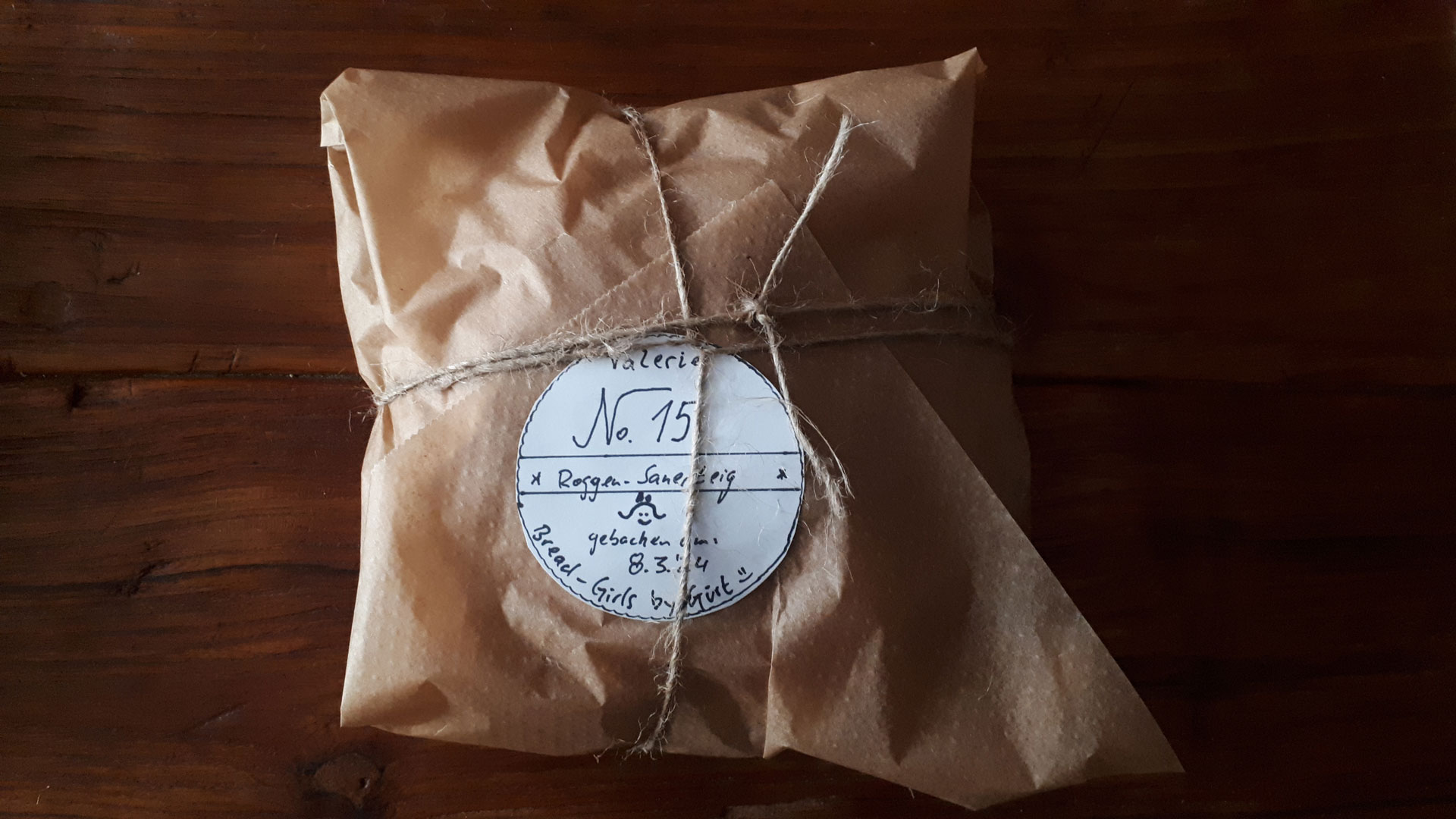 Valerie: Sauerteig-Brotlaib verpackt in Backpapier inklusive Etikett und Schleife. Auf dunkelbraunem Holztisch.